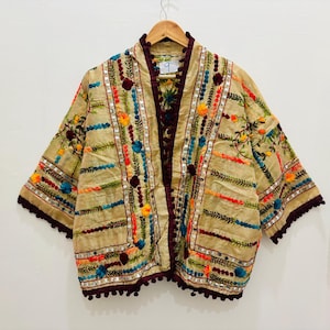 Puede incluir: Chaqueta beige bordada con coloridos estampados florales y geométricos. La chaqueta presenta un ribete burdeos oscuro, mangas cortas y está adornada con pompones. Cuelga de una percha de madera sobre un fondo blanco.