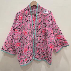 Könnte beinhalten: Rosa bestickte Jacke mit Blumen- und Rankenmustern. Die Jacke hat eine hellblaue Paspel an den Kanten und eine passende Paspel an der offenen Vorderseite. Die Stickerei ist in Rosa-, Korallen- und Schwarztönen gehalten. Die Jacke hängt an einem Holzbügel.