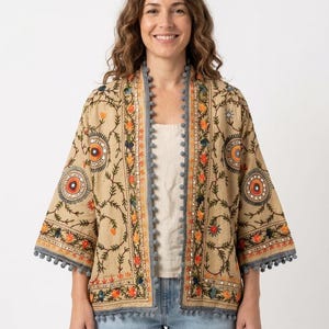 May include: A beige open-front jacket featuring intricate multi-colored floral embroidery, circular patterns with central mirrors, and grey pom-pom trim along the edges and sleeves. The garment has three-quarter length sleeves and a straight hem.