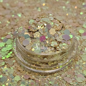 May include: A clear plastic container filled with iridescent glitter in various shapes and sizes. The glitter has a mix of colors, including gold, pink, and green. The container is sitting on a surface covered in the same glitter.