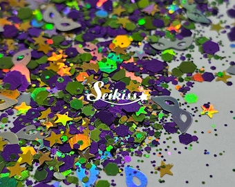 May include: A mix of green, purple, gold, and silver glitter with star and mask shapes. The glitter is scattered on a white surface.