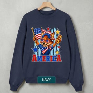 May include: A navy blue crewneck sweatshirt featuring a graphic of a fox wearing a star-spangled bandana, cap, and star-shaped sunglasses. The fox holds an American flag and a hot dog. The word AMERICA is printed in red and blue below the graphic.
