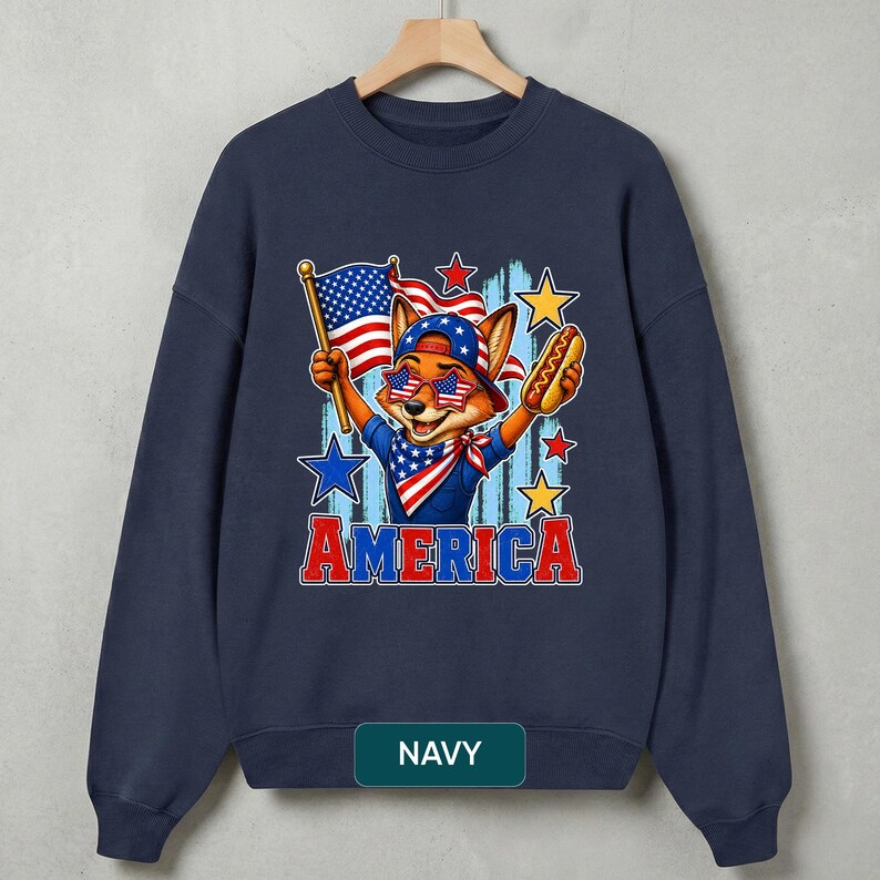 May include: A navy blue crewneck sweatshirt featuring a graphic of a fox wearing a star-spangled bandana, cap, and star-shaped sunglasses. The fox holds an American flag and a hot dog. The word AMERICA is printed in red and blue below the graphic.
