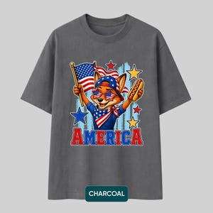May include: A charcoal gray t-shirt featuring a graphic of a fox wearing star-spangled sunglasses, a bandana, and a cap. The fox holds an American flag and a hot dog. The word AMERICA is printed in red and blue below the illustration with stars.