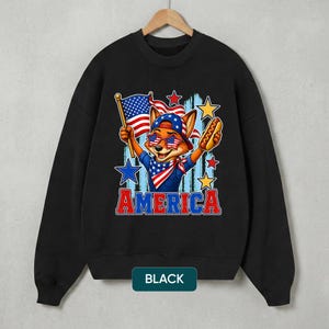 May include: A black crewneck sweatshirt featuring a graphic of a fox wearing a baseball cap and star-spangled sunglasses. The fox holds an American flag and a hot dog. The word AMERICA is printed in red and blue below the illustration with stars.