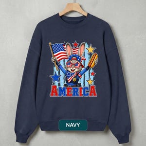 Puede incluir: Una sudadera azul marino de cuello redondo con el gr&aacute;fico de un conejo de dibujos animados que lleva una gorra, gafas de sol en forma de estrella y un pa&ntilde;uelo con motivos de bandera. El conejo sostiene una bandera de EE. UU. y un perrito caliente.