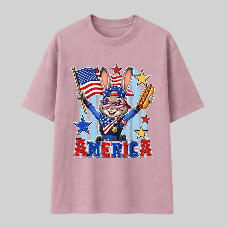 Puede incluir: Una camiseta rosa polvoriento con un conejo de dibujos animados que lleva un uniforme de polic&iacute;a, un pa&ntilde;uelo con patr&oacute;n de bandera y gafas de sol en forma de estrella. El conejo sostiene una bandera estadounidense y un perrito caliente. AMERICA en rojo y azul.