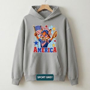 May include: A sport grey hooded sweatshirt featuring a graphic of a fox wearing star-spangled sunglasses and a bandana, holding an American flag and a hot dog. The word AMERICA is printed in red and blue block letters below the fox illustration.