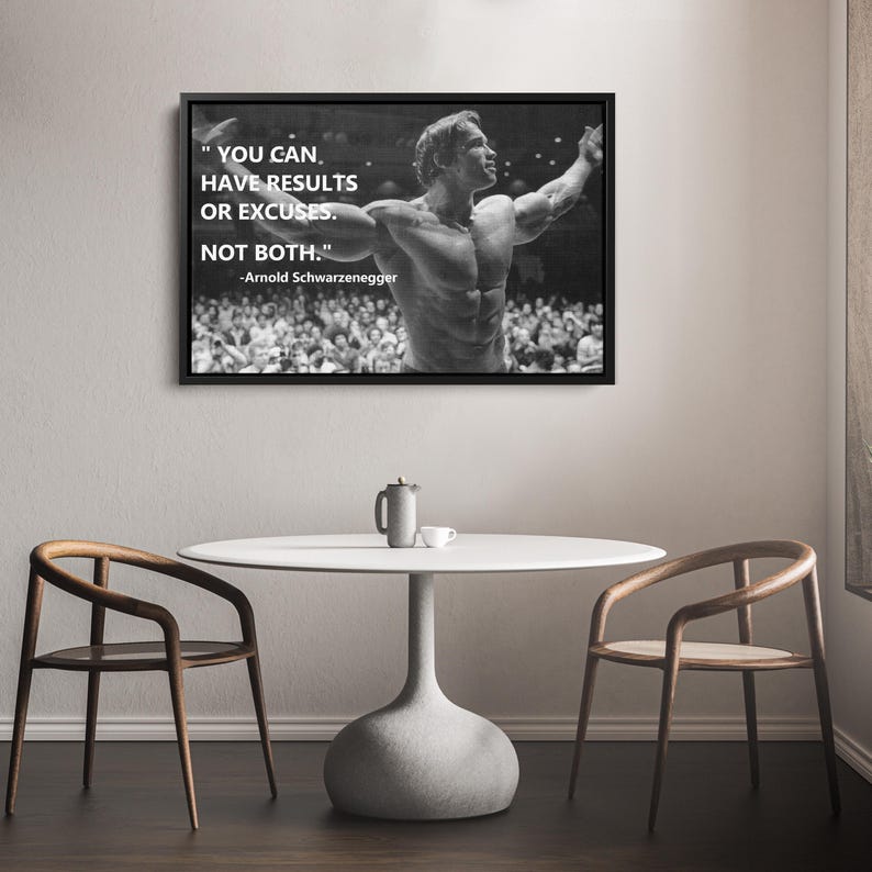 May include: Black and white framed print featuring Arnold Schwarzenegger with the quote "YOU CAN HAVE RESULTS OR EXCUSES. NOT BOTH." The image shows a bodybuilder with his arms raised, surrounded by a crowd. The print hangs above a white table and two wooden chairs.