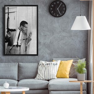 Op de afbeelding: Zwart-wit ingelijste print van een man in pak met een geweer, die uit een raam kijkt. De print hangt aan een grijze muur boven een grijze bank met kussens. Een zwarte klok en een witte hanglamp zijn ook zichtbaar.
