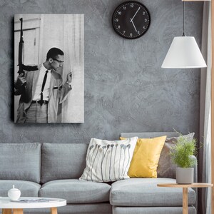Op de afbeelding: Zwart-wit wandkunst met een man in pak die een geweer vasthoudt. De afbeelding hangt aan een grijze muur boven een grijze bank met kussens. Een zwarte klok en een witte lamp zijn ook zichtbaar.