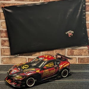 May include: A detailed, red and black toy race car with yellow accents and the text "Keep Drifting Fun!" sits on a track. A black banner with a logo is in the background.