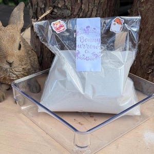 Puede incluir: Una bolsa de plástico transparente llena de un polvo de color claro, sellada con una etiqueta que dice "Bonny Burrow Co." y decorada con pegatinas. La bolsa está en un recipiente de plástico transparente, con una figura de conejo en el fondo.