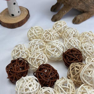 May include: A selection of woven rattan balls in cream and brown hues. The balls are arranged in a group, with a few darker brown balls mixed with the lighter ones. A decorative mushroom and a brown rabbit figurine are in the background.