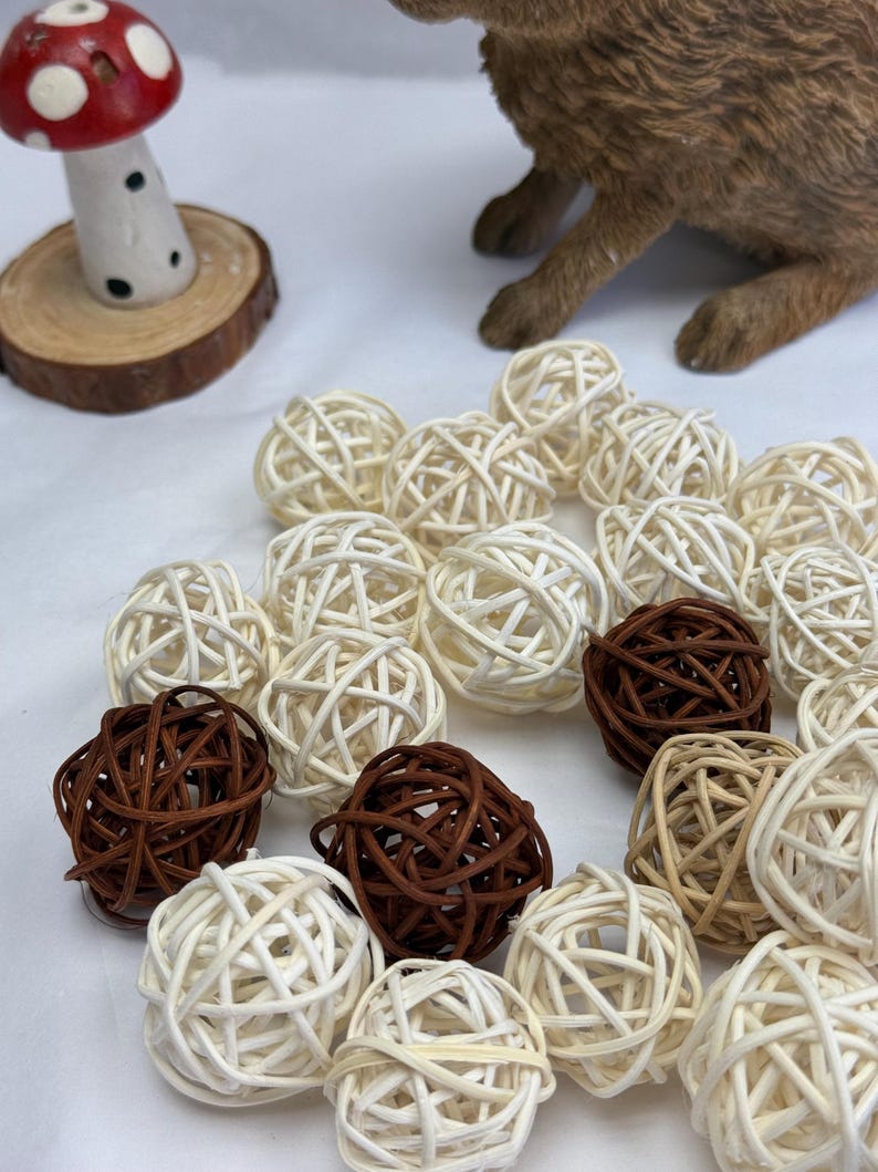 May include: A selection of woven rattan balls in cream and brown hues. The balls are arranged in a group, with a few darker brown balls mixed with the lighter ones. A decorative mushroom and a brown rabbit figurine are in the background.