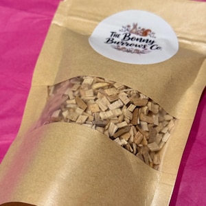 Puede incluir: Una bolsa de papel kraft con una ventana transparente, llena de pequeñas virutas de madera. Una pegatina ovalada blanca con el texto "The Bonny Burrows Co." está en la parte delantera. El fondo es una superficie rosa brillante.