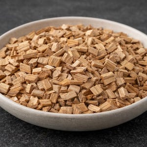 Puede incluir: Un cuenco de cerámica blanca lleno de virutas de madera de color marrón claro. Las virutas son pequeños trozos rectangulares, probablemente para ahumar o asar a la parrilla. El cuenco está sobre una superficie gris oscuro.