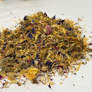 May include: A pile of dried flowers and petals in shades of yellow, blue, pink, and brown. The mix includes whole flower heads and fragmented pieces, creating a textured, natural appearance. The image is a close-up shot, highlighting the details of the botanical elements.