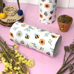 Op de afbeelding: Witte cilindrische papieren buizen versierd met zonnebloemen, bijen en groene bladeren. Eén buis is horizontaal, de andere verticaal. Een kleine terracotta pot met een geschilderde bij en mos staat in de buurt. Gedroogde gele bloemen en een houten ringstandaard zijn ook aanwezig.