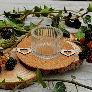 Może przedstawiać: Przezroczysty, żebrowany szklany świecznik na tealight stoi na okrągłej podstawce korkowej z dwoma drewnianymi wycięciami w kształcie serca. Podstawka spoczywa na drewnianym plastrze, otoczonym świeżymi jeżynami i zielonym bluszczem.