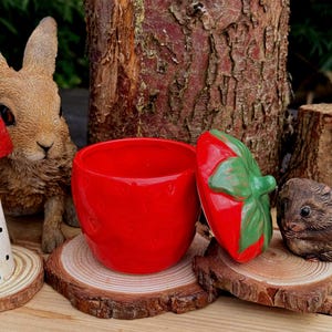 May include: A vibrant red ceramic strawberry-shaped pot with a matching lid. The lid is green and red, and the pot sits on a wooden slice. A rabbit and a small animal figurine are in the background.