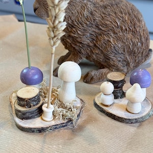 May include: A whimsical display featuring wooden mushroom figurines, purple spheres, and small log slices arranged on wood rounds. A brown rabbit figurine is in the background. A sprig of wheat and a green stem with a purple sphere add to the rustic charm.