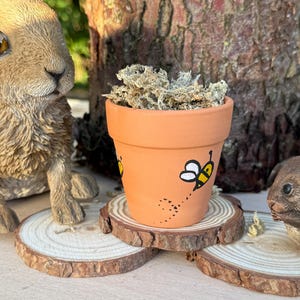 Op de afbeelding: Een kleine bloempot in terracotta kleur met een handgeschilderd bijenontwerp. De pot is gevuld met gedroogd mos en staat op een houten schijf. Een decoratief konijnenbeeld is zichtbaar op de achtergrond.