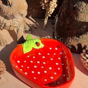 May include: A bright red ceramic strawberry-shaped dish with white spots. The dish features a green leaf and a small ladybird detail. This decorative item could be used for serving snacks or as a trinket tray.
