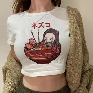 Può includere: Top corto bianco con un'illustrazione a cartoni animati di un personaggio in una ciotola di ramen, con testo giapponese sopra. Il personaggio ha i capelli rosa e indossa un kimono rosa. La ciotola di ramen è rossa con un design decorativo.