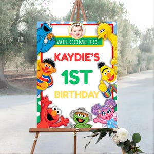 以下が含まれることがあります： セサミストリートのキャラクターが描かれた、カラフルな誕生日サイン。「Kaydie's 1st Birthday」と書かれており、赤ちゃんの写真が添えられています。クッキーモンスター、エルモ、ビッグバードなど、他のキャラクターがテキストを囲んでいます。