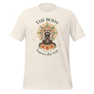 May include: Cream-colored t-shirt featuring a raccoon in a meditative pose, surrounded by a floral mandala design. The text "THE BODY knows the way" is arched above and below the raccoon illustration. The shirt is made of a soft material.