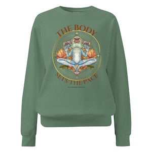 Könnte beinhalten: Salbeigrünes Sweatshirt mit einer Grafik einer meditierenden Froschfigur in Lotusposition, umgeben von einem goldenen Kreisdesign und dem Text "THE BODY SETS THE PACE". Das Design enthält orangefarbene und blaue Akzente.