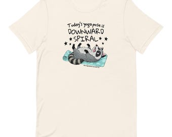 May include: Cream-colored t-shirt featuring a cartoon raccoon lying on a yoga mat. The raccoon is in a relaxed pose, with the text "Today's yoga pose is DOWNWARD SPIRAL" above it. The shirt is made of a soft material.