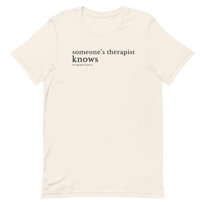 Pode incluir: Camiseta creme com o texto preto "someone's therapist knows" e "the regulated raccoon co." abaixo. A camiseta tem mangas curtas e gola redonda.