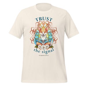 以下が含まれることがあります： クリーム色のTシャツ。ステンドグラスのデザインの中にキツネのカラフルなイラストが描かれています。「TRUST the signal」の文字がキツネの上と下にアーチ状に配置されています。「the regulated raccoon co.」のロゴが下にあります。