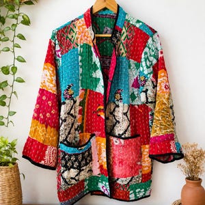 May include: A long-sleeved jacket featuring a patchwork design of various colorful fabrics in shades of red, green, blue, and yellow. The garment includes two front pockets, a black trim along the edges, and visible white stitching throughout.