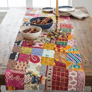 Può includere: Un runner da tavola rettangolare caratterizzato da un design patchwork composto da vari quadrati di tessuto nei toni del rosso, giallo, blu e bianco. Il tessuto presenta diversi motivi tra cui stampe paisley, floreali e geometriche.