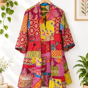 Pode incluir: Um vestido de manga comprida com um design de patchwork vibrante, apresentando vários padrões florais e geométricos em tons de vermelho, amarelo, rosa, verde e azul. A peça tem um decote em V, uma saia em camadas e costuras brancas visíveis.