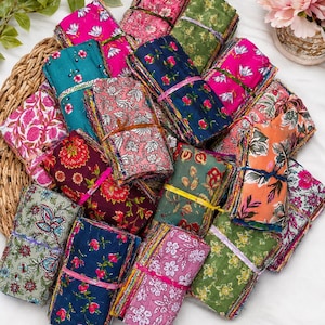 May include: A collection of fabric bundles featuring various floral and botanical prints in vibrant colours including pink, teal, navy blue, green, and orange. Each bundle is tied with a thin string and arranged in a cluster on a woven surface.