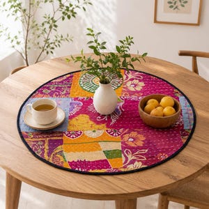 May include: A round, quilted fabric placemat featuring a patchwork design of vibrant pink, orange, and blue floral patterns with visible white stitching. The placemat is centered on a wooden table and holds a white ceramic vase, a cup, and a bowl.