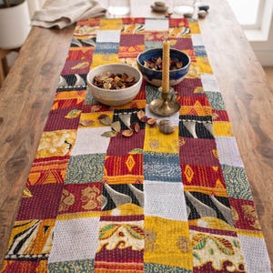 May include: A rectangular patchwork table runner featuring a variety of fabric squares in shades of red, yellow, white, and blue. The fabric displays diverse patterns including floral, geometric, and abstract motifs, all joined by visible white stitching.