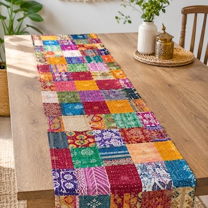 Puede incluir: Un camino de mesa rectangular con un diseño de patchwork compuesto por numerosas piezas de tela cuadradas pequeñas en colores vibrantes que incluyen magenta, verde azulado, amarillo, naranja y rojo intenso, unidas con detalles de costura blanca.