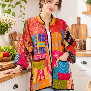 May include: A long-sleeved, open-front jacket featuring a vibrant patchwork design with various colourful patterns in shades of red, orange, yellow, blue, and green. The garment includes a mandarin collar, black trim along the edges, and two pockets.