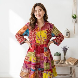 Op de afbeelding: Een knielange jurk met een levendig patchworkontwerp met diverse bloemmotieven en geometrische patronen in tinten rood, geel, paars en blauw. Het kledingstuk heeft een opstaande kraag, een knoopsluiting aan de voorzijde en driekwartmouwen.