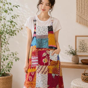 Puede incluir: Un delantal sin mangas con un diseño de patchwork que presenta varios cuadrados de tela coloridos en tonos rojos, amarillos, azules y blancos. El delantal tiene un escote cuadrado, tirantes ajustables con hebillas y un bolsillo frontal derecho.