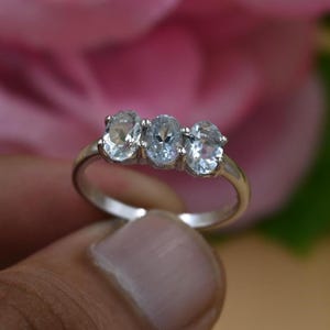May include: A silver ring with three oval, pale blue gemstones. The ring is set against a blurred pink and green background. The gemstones are the focus, highlighting their clarity and colour. The ring is a piece of jewellery.