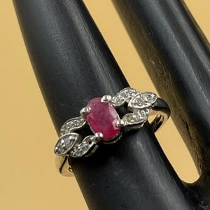 Peut inclure: Bague en argent ornée d'un rubis ovale rouge au centre, entouré de petits diamants clairs. La bague présente un motif décoratif en forme de feuilles de chaque côté de la pierre centrale. La bague est présentée sur une surface noire.