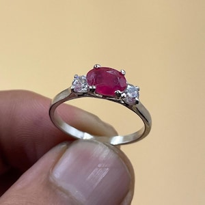 Puede incluir: Un anillo de plata con un gran rubí ovalado, flanqueado por dos piedras preciosas claras y redondas más pequeñas. El rubí es de color rojo vibrante, y el anillo está sostenido por los dedos de una persona sobre un fondo beige claro.