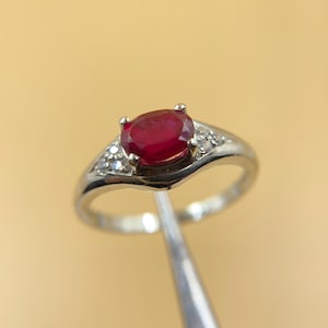 Peut inclure: Une bague en argent ornée d'une grosse pierre rouge foncé de forme ovale en son centre. De petites pierres claires sont serties de chaque côté de la pierre principale. La bague est tenue par un outil argenté sur fond beige clair.