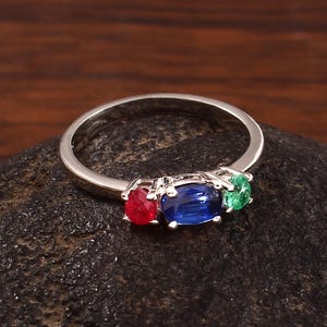 Può includere: Un anello d'argento con tre pietre preziose ovali: rossa, blu e verde. Le pietre sono disposte in fila sulla fascia dell'anello. L'anello è esposto su una superficie scura e strutturata.